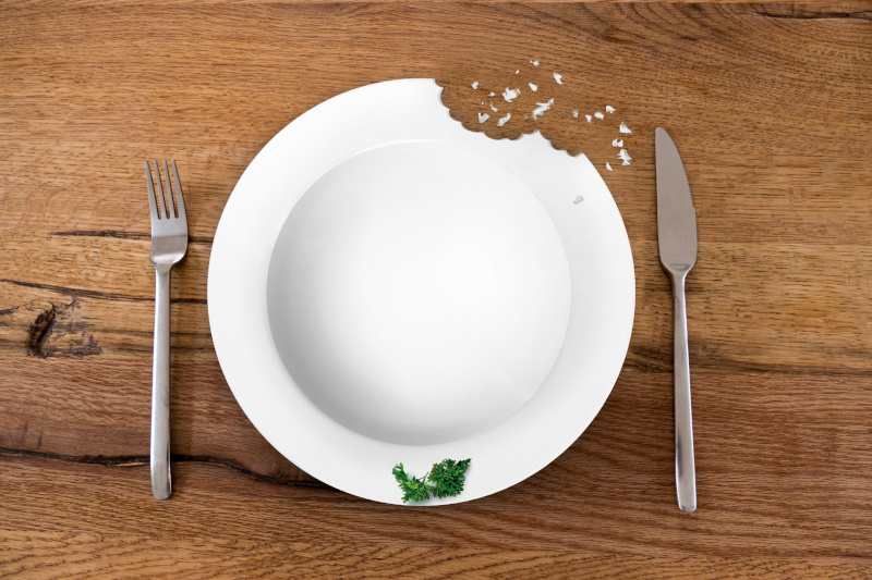 Das Bild zeigt einen weißen Teller zwischen Messer und Gabel auf einem Tisch. Der Teller ist außen angeknabbert und leer, nur auf dem Rand liegt ein kleines Blatt Petersilie.